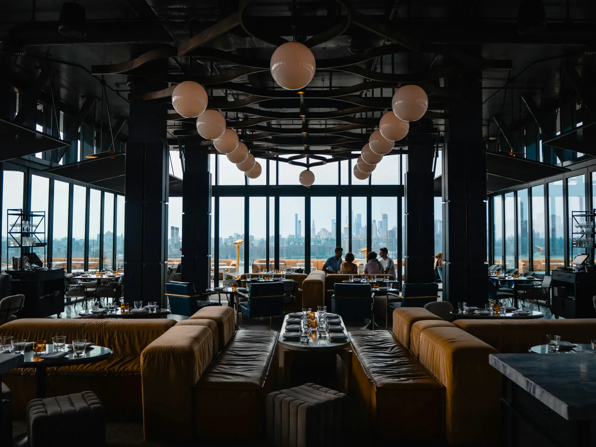 Dark, atmospheric rooftop restaurant interior with velvet banquette seating, globe pendant lighting, and a city skyline view