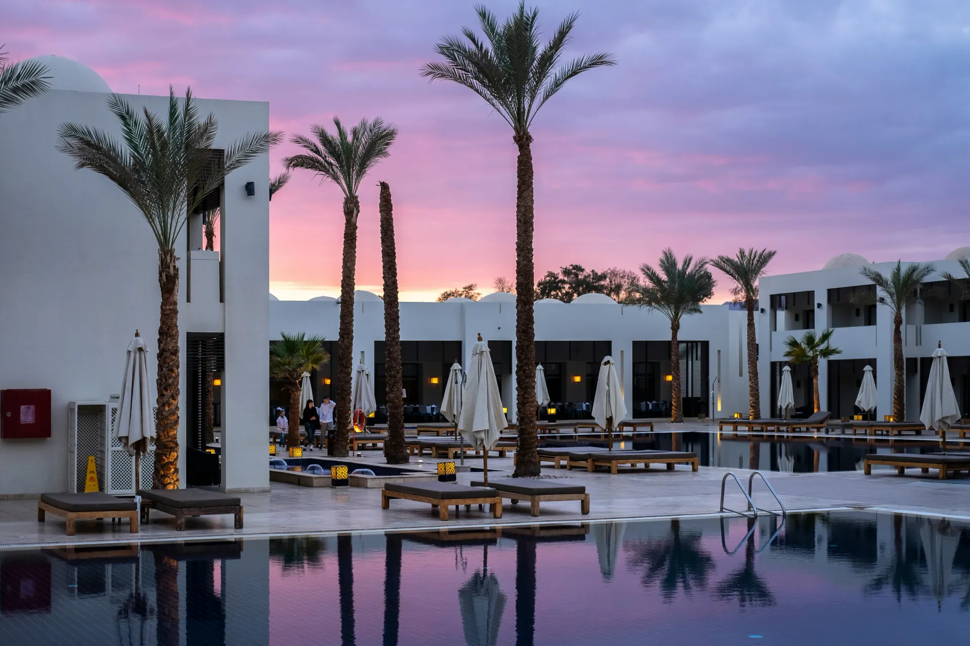 Resort pool deck and outdoor lounge area at dusk, framed by palm trees and Mediterranean-style architecture