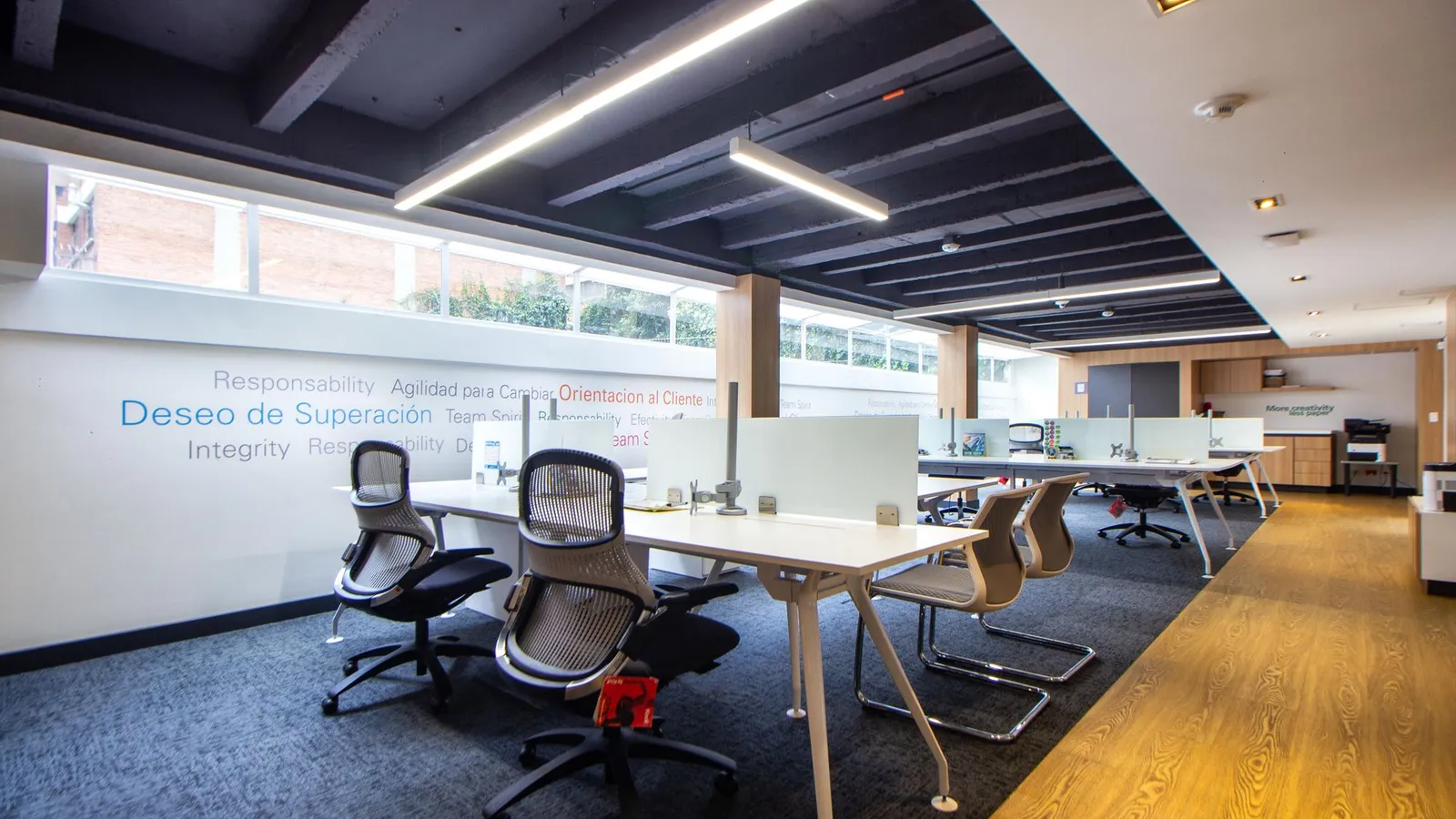 Long open-plan office with white desks, mesh chairs, linear ceiling lights and a wood and carpet floor