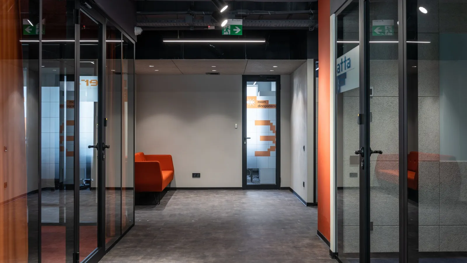 Office corridor with black-framed glass partitions, a glazed door and an orange sofa beyond