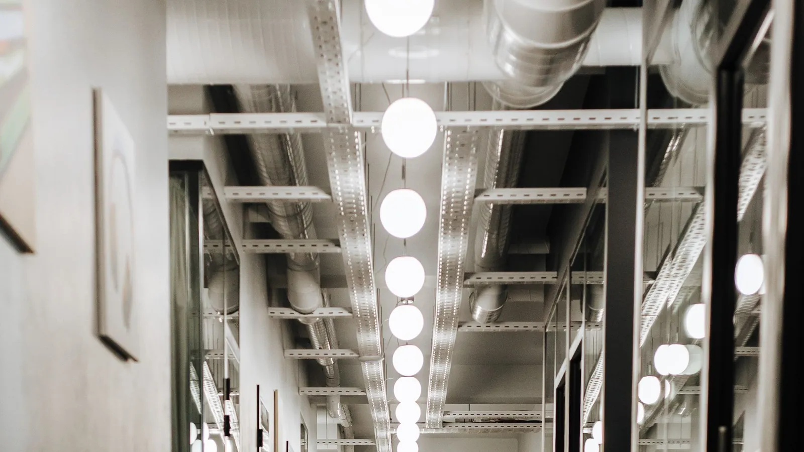 Ceiling void with perforated cable trays, ducts and a row of globe lights above a glazed office corridor