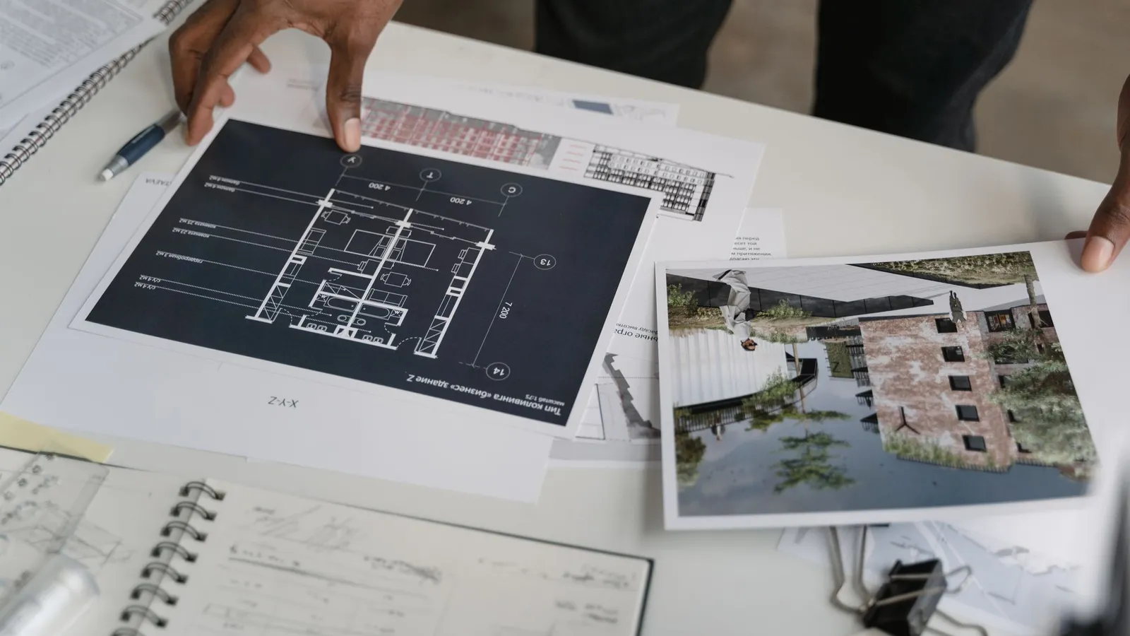 Hands resting on a printed floor plan beside a site photograph and a spiral notebook
