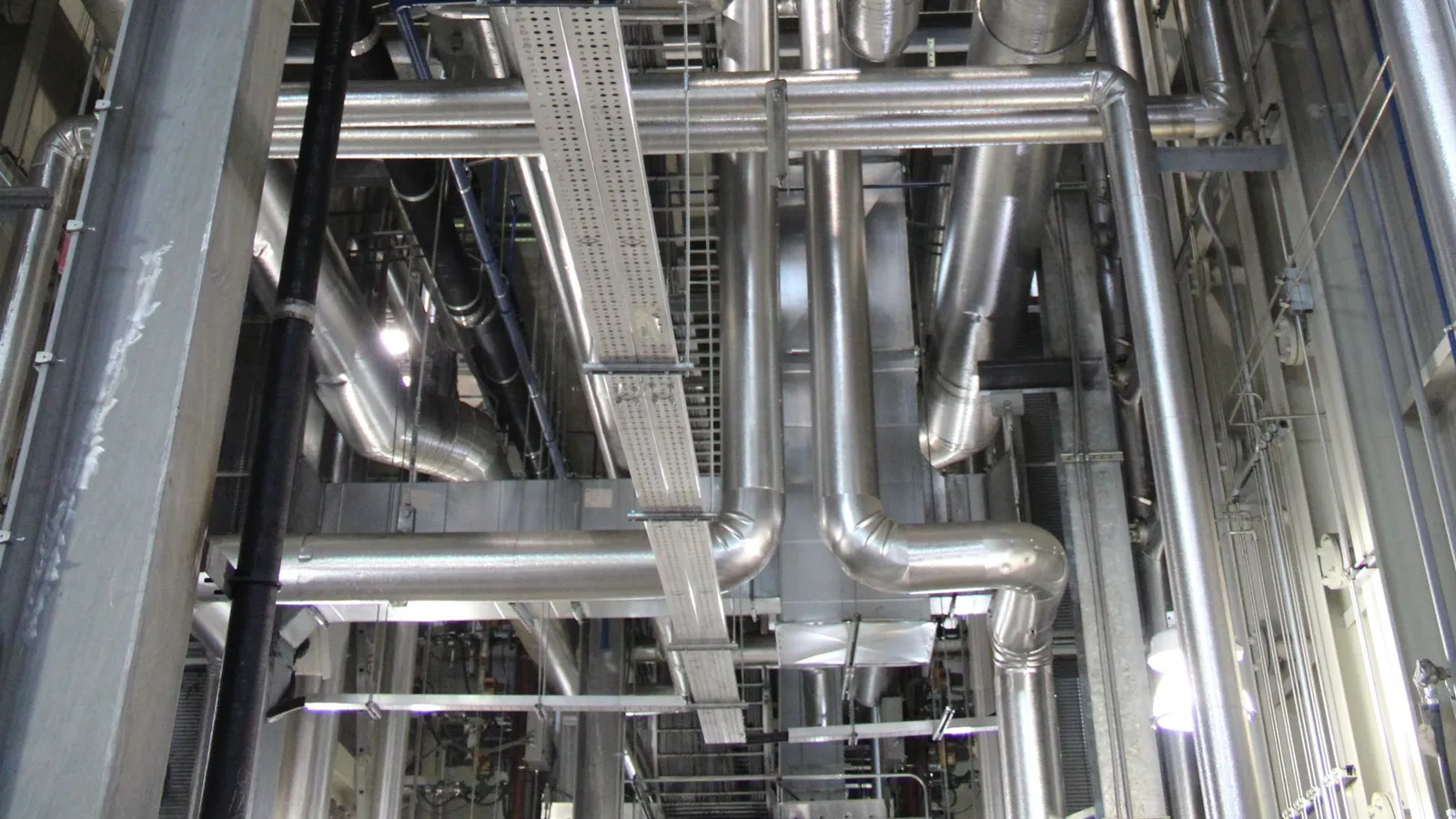 Polished steel ducts and pipework running beside a perforated cable tray in a plant area