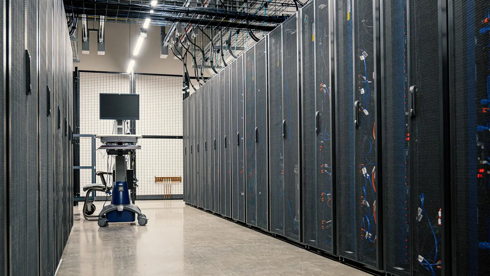 Aisle of dark server cabinets with overhead cable trays leading to a monitor cart in a bright data hall