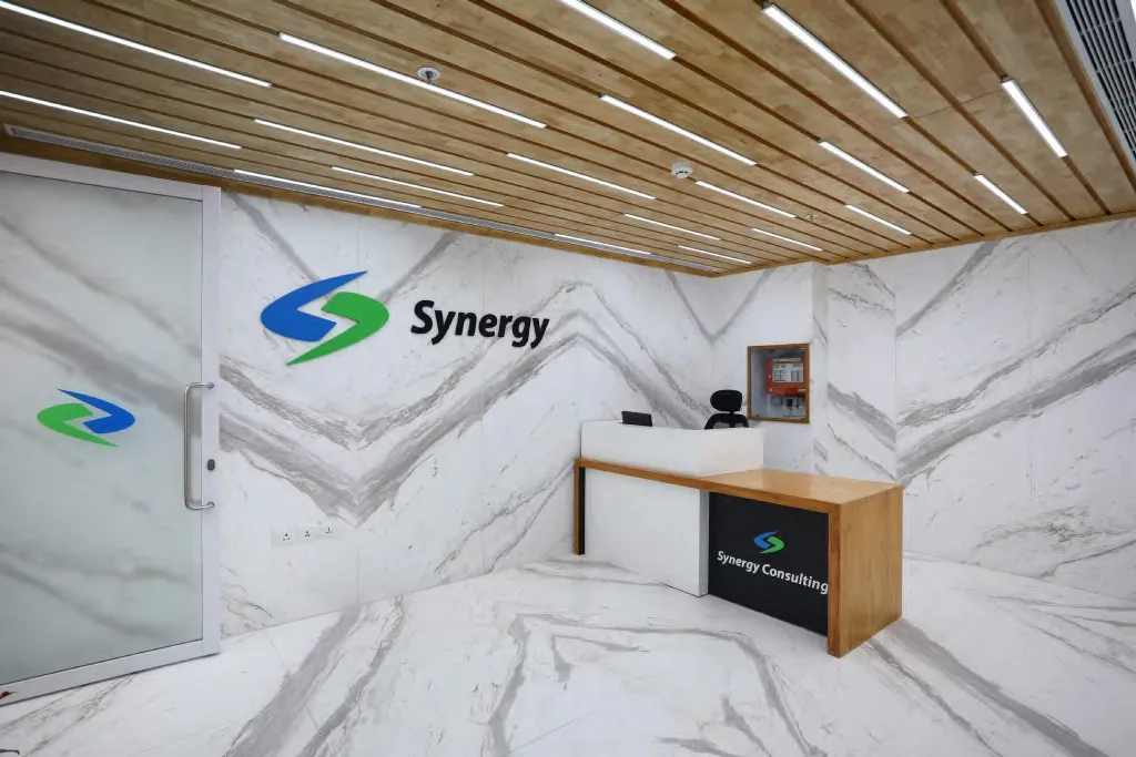 Synergy Consulting reception in Jasola, New Delhi, with marble-look wall panels, a timber slat ceiling and a timber reception desk
